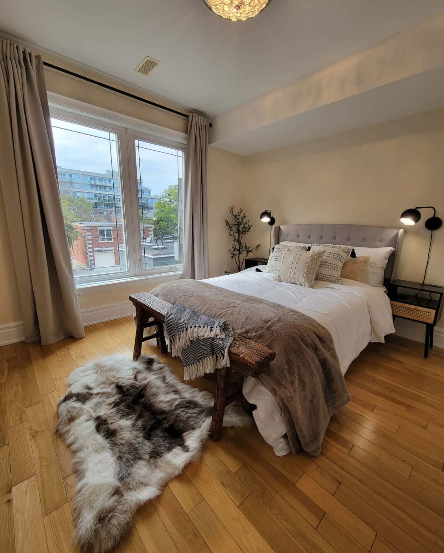 A stylish urban bedroom with warm honey hardwood floors and soft beige walls. A queen-size bed with a gray tufted wingback headboard is dressed in white linens, a taupe faux-fur throw, and textured neutral accent pillows. A rustic wooden bench at the foot of the bed holds a folded plaid blanket. A gray-and-white cowhide rug lies on the floor in the foreground. Floor-to-ceiling windows with taupe linen curtains reveal a city street view with brick buildings and green trees. Two black wall-mounted reading lamps flank the bed, and a faux olive branch in a vase adds a natural touch. A warm globe pendant light hangs from the white ceiling.