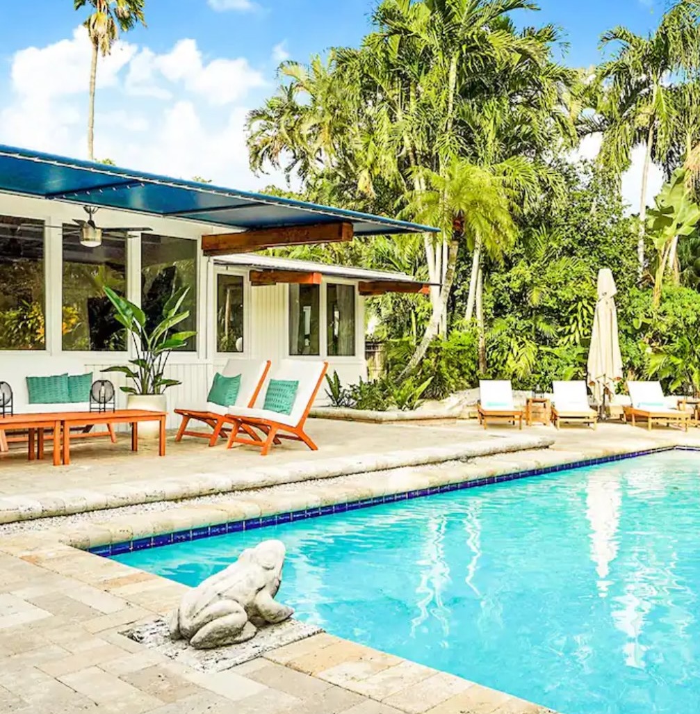 Tropical Airbnb poolside patio with lounge chairs, palm trees, modern outdoor seating, and a private pool beside a bright vacation home.