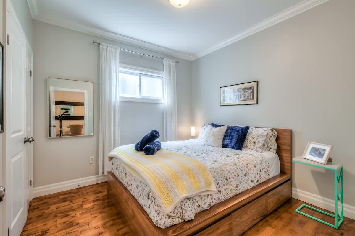 A cozy guest bedroom with light gray walls and warm hardwood flooring, featuring a queen-size bed on a modern walnut platform frame. The bed is dressed with a white floral duvet, a yellow-and-white striped knit throw, navy blue rolled towels, and a navy velvet accent pillow. Sheer white curtains frame a single window, with a small glowing table lamp beside the bed and a framed art print on the wall above.