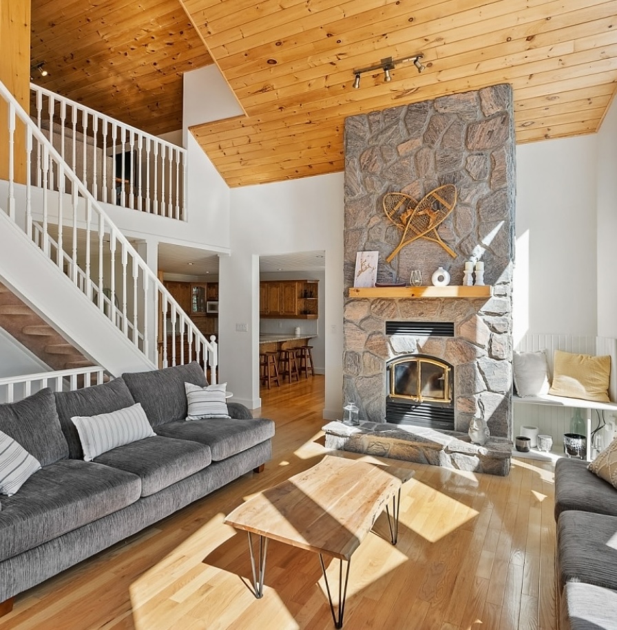 Bright Airbnb cabin living room with vaulted wood ceilings, stone fireplace, gray sofas, open staircase, and natural light.