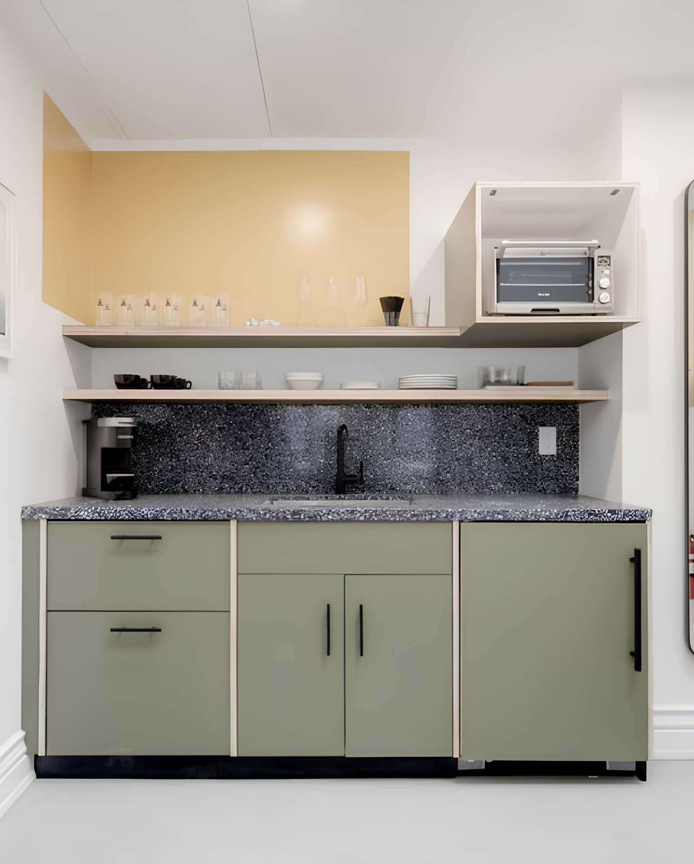 A compact kitchenette with sage green flat-panel cabinetry and black bar handles, set against white walls. A blue-gray speckled granite countertop and matching backsplash surround a single black faucet sink. A black drip coffee maker sits on the left counter. Above, a light wood floating shelf holds clear wine glasses, tumblers, and white dishes. A warm butter-yellow accent wall fills the upper portion behind a dropped ceiling soffit. In the upper right, a white wall-mounted cabinet houses a small stainless steel toaster oven. The floor is dark, and white baseboards trim the bottom of the walls.