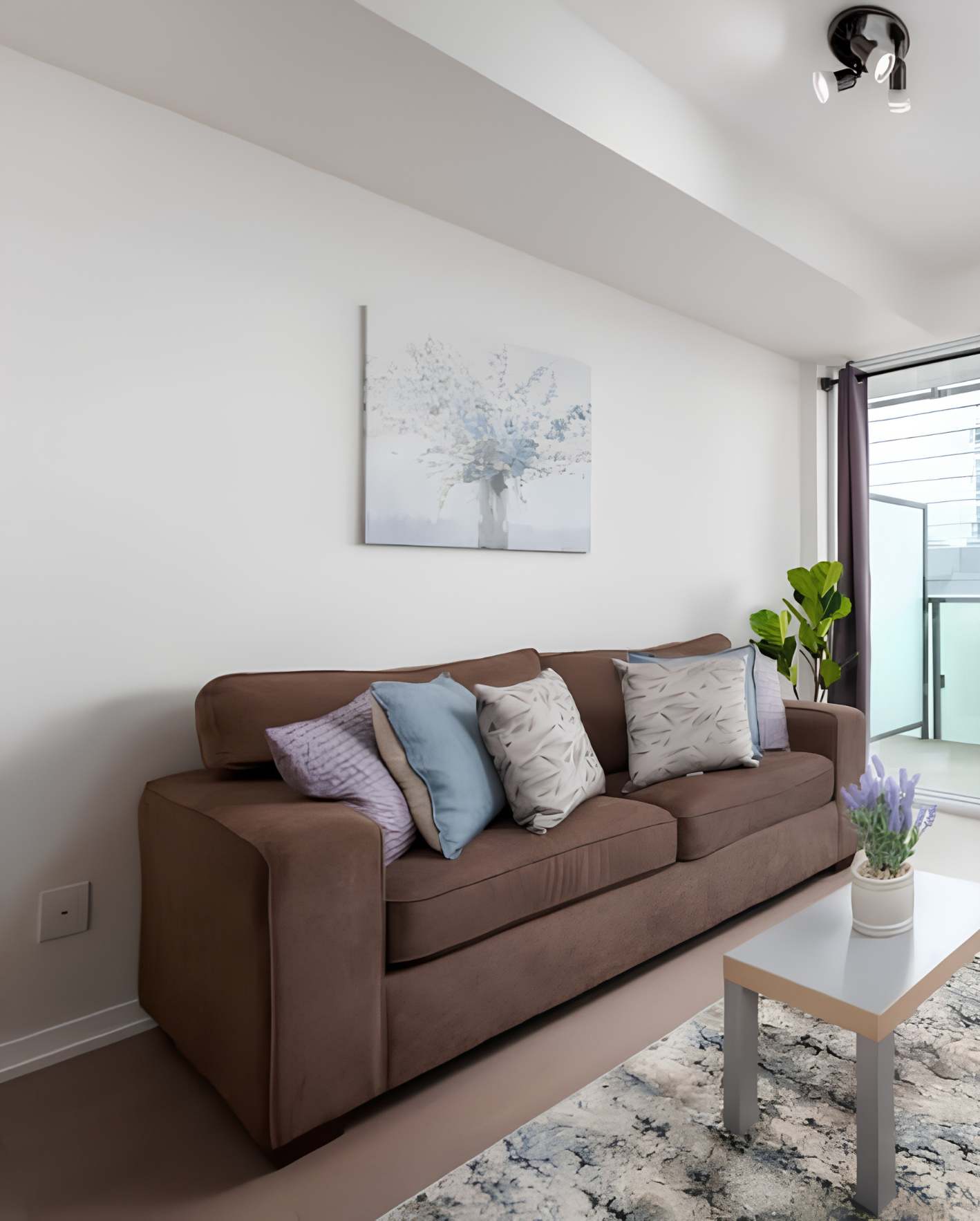 A compact modern living room with white walls and a brown microfiber three-seat sofa with pastel accent pillows in blue, lavender, and a gray leaf pattern. Above the sofa hangs a soft blue floral canvas print in a vase. A green fiddle-leaf fig plant sits beside a sliding glass balcony door with dark curtains. In the foreground, a white-and-wood coffee table holds a small pot of lavender flowers, set on a blue-and-white marbled area rug. A black modern ceiling spotlight is mounted above.