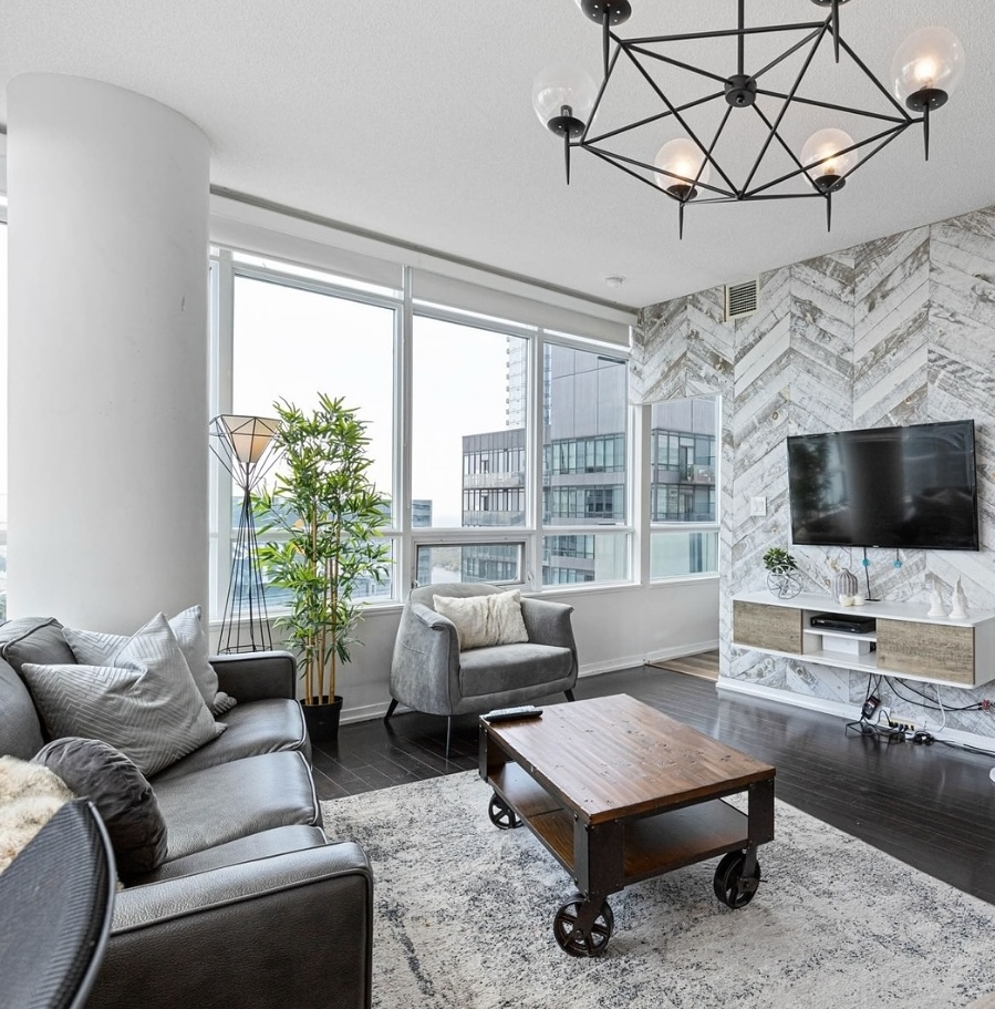 Modern Airbnb living room with leather sofa, accent chair, large windows, wall-mounted TV, wood coffee table, and contemporary city condo decor.
