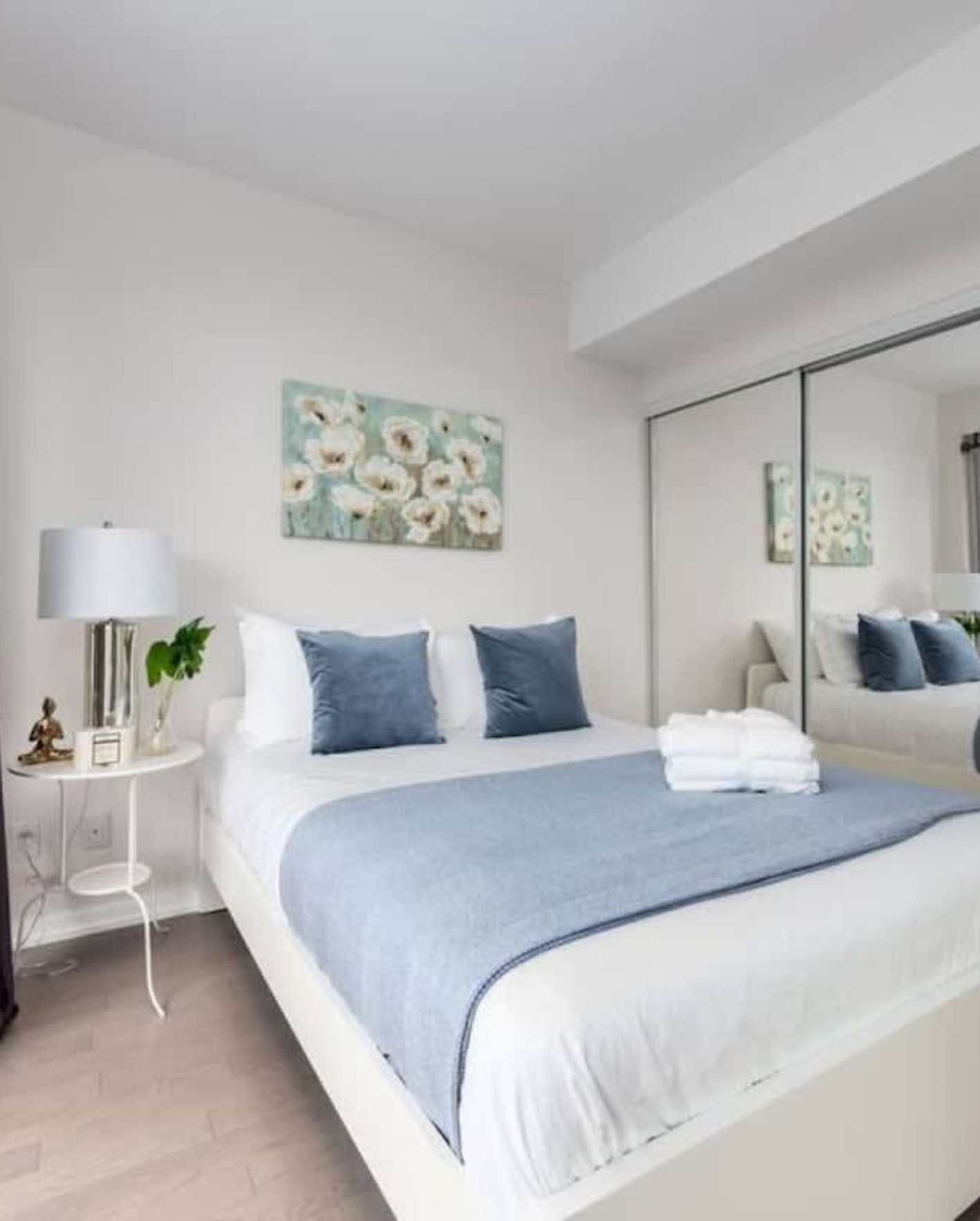 A bright, modern guest bedroom with white walls and light herringbone wood flooring. A queen-size bed with a white platform frame is dressed in crisp white linens, a dusty blue throw blanket, navy blue accent pillows, and neatly folded white towels. Above the bed hangs a teal and white floral canvas painting. A white round nightstand holds a glass table lamp, a small green plant, and a brass Buddha figurine. A large mirrored sliding closet door on the right reflects the room, creating a sense of added space.