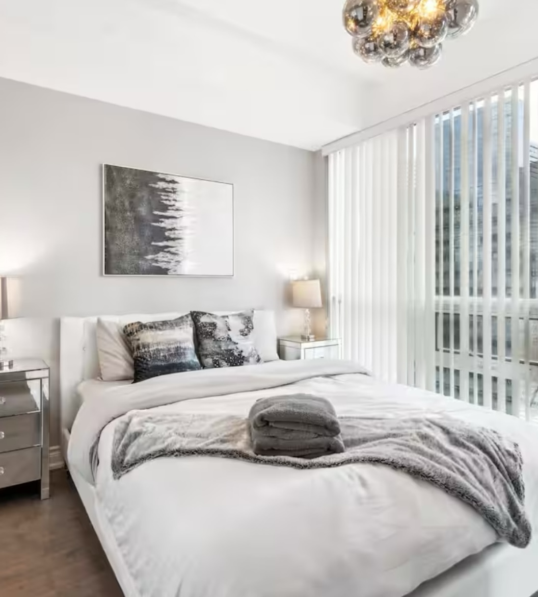 Bright Airbnb bedroom with white bedding, mirrored nightstands, folded towels, abstract wall art, and floor-to-ceiling windows with city views.