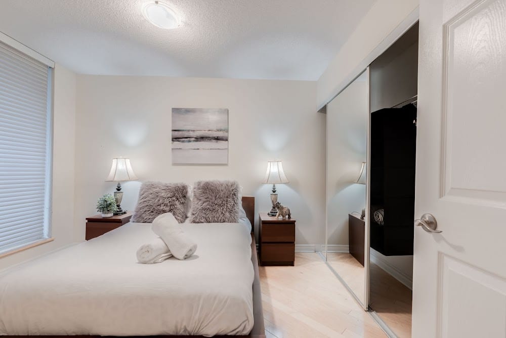 Cozy Airbnb bedroom with white bedding, rolled towels, matching bedside lamps, mirrored closet doors, and soft neutral decor.
