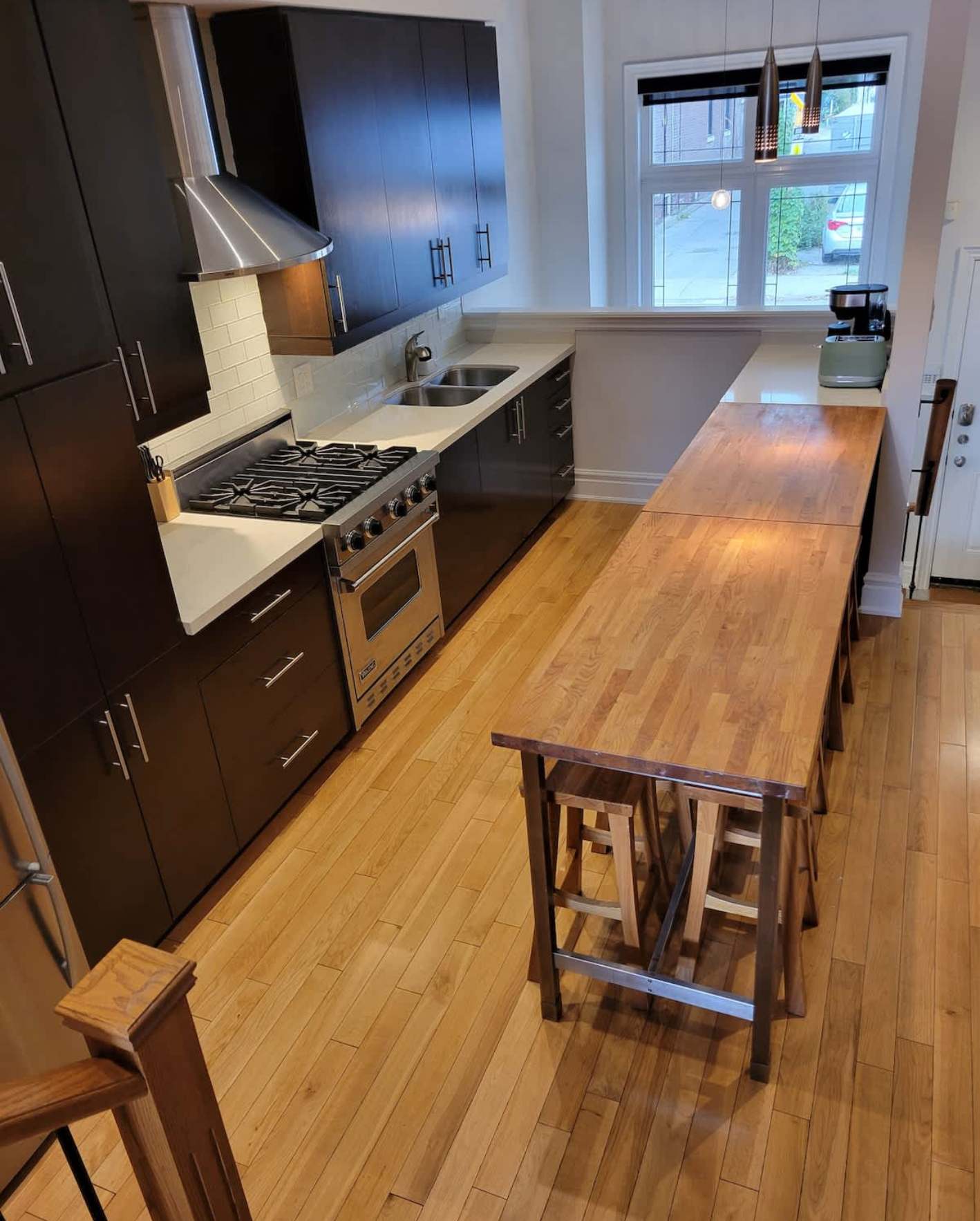 A narrow galley kitchen with warm honey hardwood flooring and white walls. Dark espresso-brown flat-panel cabinetry lines the left wall with stainless steel bar handles, a white quartz countertop, white subway tile backsplash, and a stainless steel gas range with a conical range hood above. A double window at the far end lets in natural light, with a black coffee maker on the counter below. On the right side, a long butcher-block countertop or bar extends the length of the room, with a rustic wooden trestle-style high table and two matching wooden bar stools tucked underneath. A wooden barstool is partially visible in the lower-left foreground.