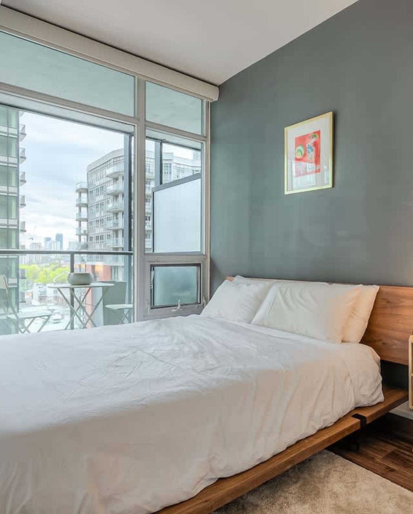 A modern high-rise bedroom with a muted blue-green accent wall and floor-to-ceiling glass windows overlooking neighboring condo buildings and treetops. A queen-size bed with a low walnut platform frame is dressed in simple white bedding with two pillows. A small colorful abstract art print in a gold frame hangs above the bed. Through the glass, a small balcony is visible with a folding bistro table and chair. Dark hardwood flooring and a white area rug complete the space.