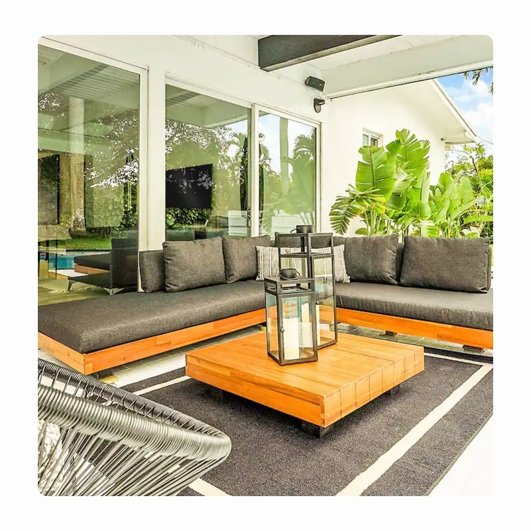 Stylish Airbnb patio with outdoor sectional seating, wood coffee table, tropical plants, and covered lounge area beside the pool.