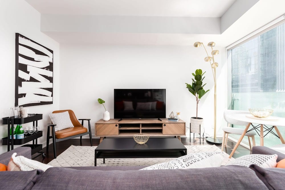 Modern Airbnb living room with neutral decor, TV console, leather accent chair, bar cart, floor lamp, indoor plants, and large windows with natural light.