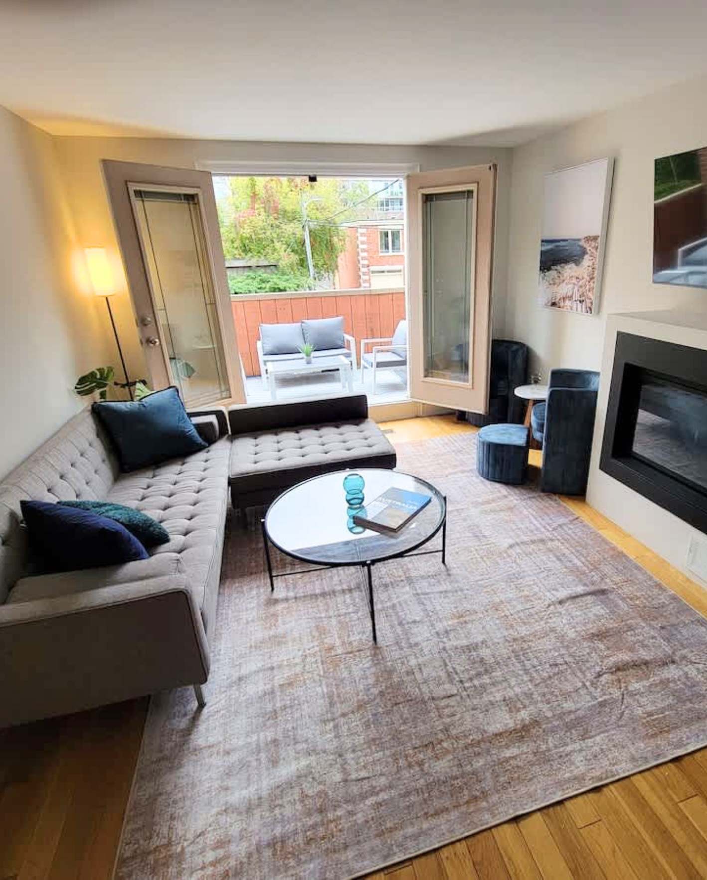 A modern ground-floor living room with warm honey hardwood floors and cream walls. A large gray tufted L-shaped sectional sofa with a matching ottoman is arranged around a round glass-top coffee table with thin black metal legs. Navy blue accent pillows and a teal throw blanket add color. A large muted pink-and-beige vintage-style area rug anchors the seating area. French doors with transom windows open to a small patio with gray outdoor furniture and a red-stained wooden fence. A tall floor lamp with a white shade stands in the corner beside a potted plant. On the right wall, a modern linear gas fireplace is built into a dark surround, with two framed art prints above—one coastal, one abstract green. A dark navy armchair and a small blue velvet ottoman sit beside a tiny round side table.