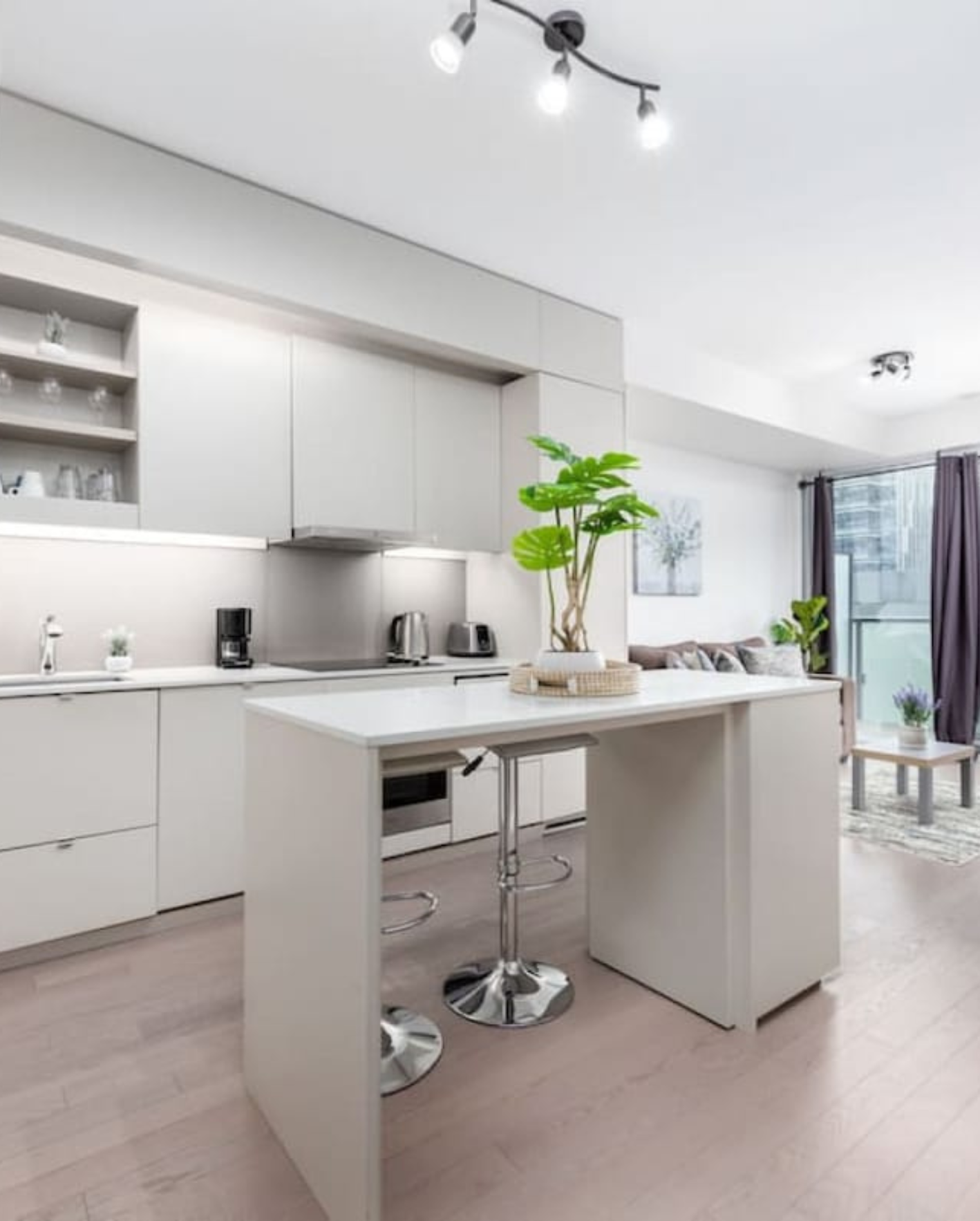 A modern open-concept kitchen and living area in a high-rise condo with light hardwood floors and white walls. The minimalist kitchen features flat-panel gray and white cabinetry, a white quartz island with two chrome adjustable bar stools, and a large tropical monstera plant in a vase as a centerpiece. A black track light fixture runs along the ceiling. In the background, a gray sofa with throw pillows faces a glass balcony door with dark curtains, offering a city view.