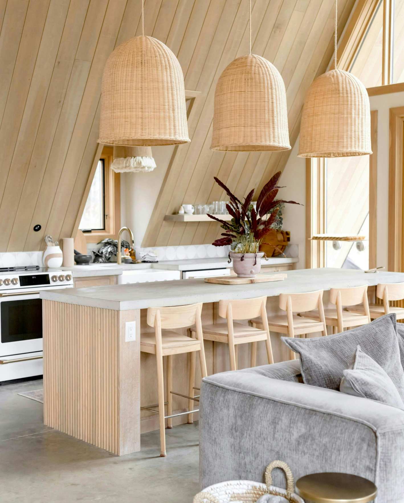 A modern Scandinavian-style kitchen with a vaulted wood-paneled ceiling, featuring three large woven rattan pendant lights hanging over a concrete-topped island with vertical wood slat detailing. Light wood bar stools line the island, and a gray upholstered armchair sits in the foreground. The space includes white appliances, open shelving, and a vase of dried burgundy foliage as an accent.