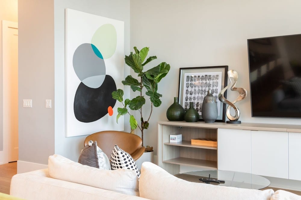 Modern Airbnb living room with abstract wall art, accent chair, indoor plants, decorative shelving, and wall-mounted TV in a bright, staged interior.