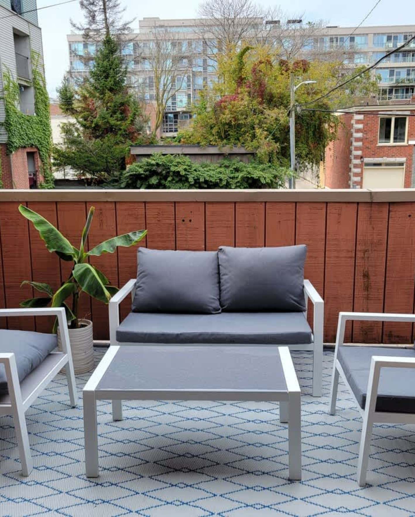 A rooftop patio seating area with a white aluminum-frame loveseat and matching armchairs, all with charcoal gray cushions, arranged around a low white rectangular coffee table. A blue-and-white diamond-pattern outdoor rug covers the deck. A potted banana leaf plant in a gray ceramic planter sits to the left. Behind the furniture, a weathered red-brown horizontal wood fence provides privacy. In the background, mature green trees, a red brick building, and a mid-rise glass condo tower are visible under a partly cloudy sky.