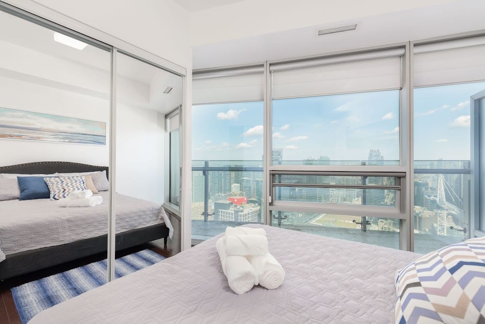 Bright Airbnb bedroom with mirrored closet doors, neatly made bed, rolled towels, and floor-to-ceiling windows overlooking a city skyline and water view.