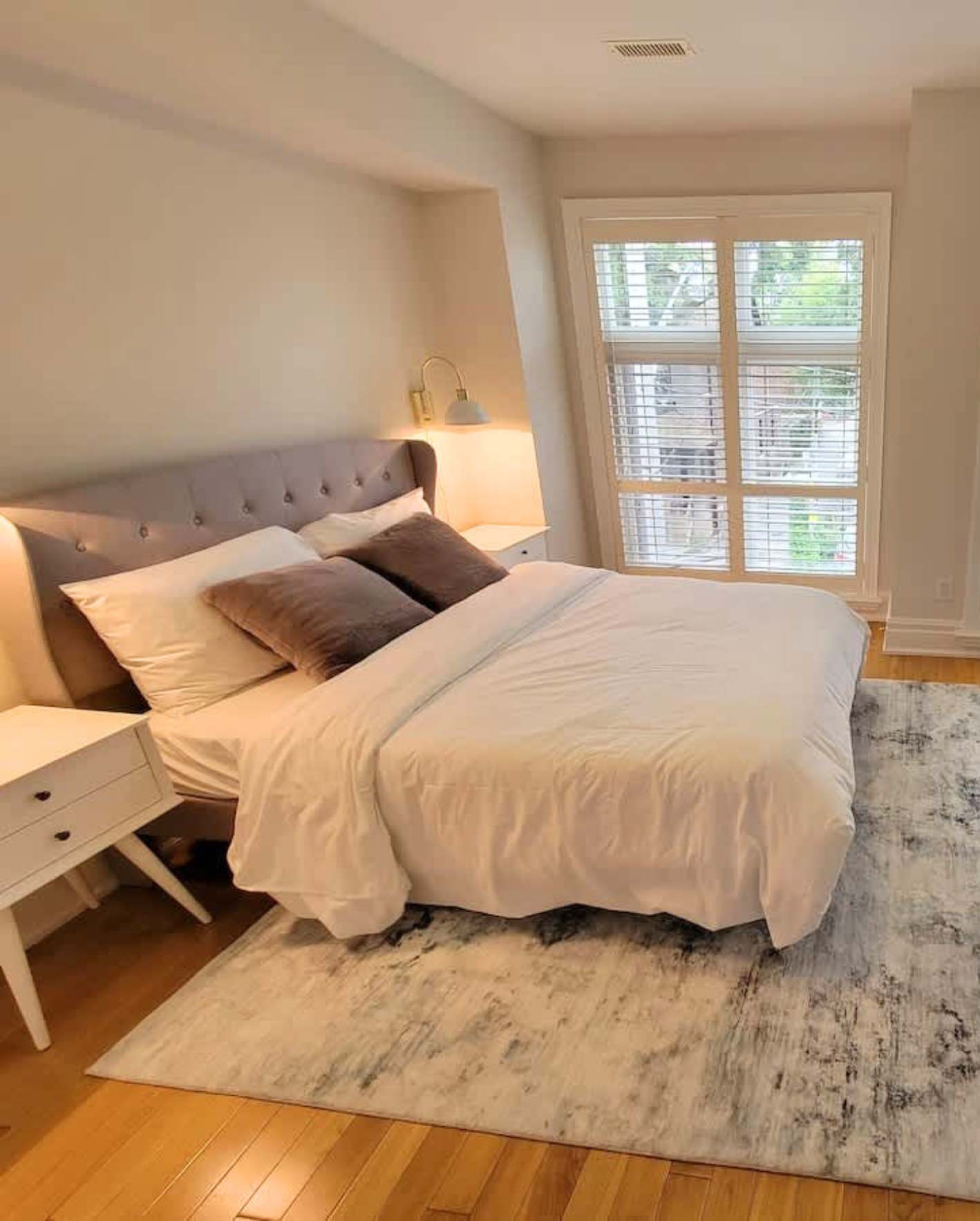 A cozy bedroom with warm beige walls and honey hardwood flooring. A king-size bed with a gray tufted wingback headboard is dressed in crisp white bedding with two mauve velvet accent pillows. Two brass wall-mounted sconces with white shades flank the headboard. A white mid-century modern nightstand sits to the left. A large window with white plantation blinds lets in natural light, revealing green foliage outside. A gray-and-white abstract marbled area rug covers the floor beside the bed. The ceiling has a slight slope on the left side, suggesting an upper-floor room.