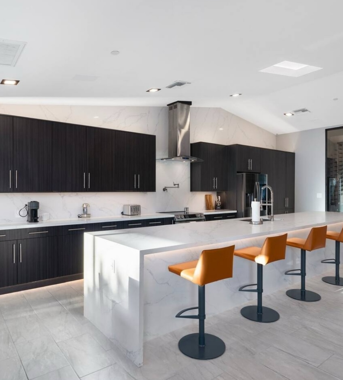 Modern Airbnb kitchen with dark wood cabinetry, oversized waterfall island, white countertops, stainless steel appliances, and orange bar stools.