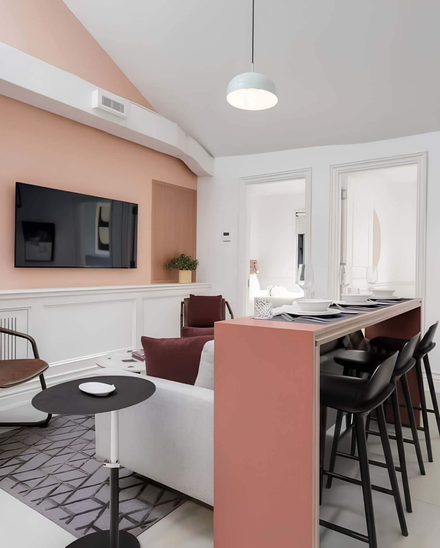 A compact open-plan living and dining area with a muted blush-pink accent wall and white wainscoting below. A wall-mounted flat-screen TV hangs on the pink wall above a small potted plant on the ledge. A white sofa with a deep burgundy accent pillow sits against the wall, with a round black bistro table and a mid-century wooden armchair to the left on a gray geometric-patterned area rug. On the right, a pink-painted kitchen island or bar counter with a light wood top is set with dark place settings and flanked by four black molded bar stools. A pale mint-green dome pendant light hangs from the white ceiling above. Two tall white-framed windows on the far wall let in natural light, and a white structural beam crosses the ceiling where it meets the pink wall.