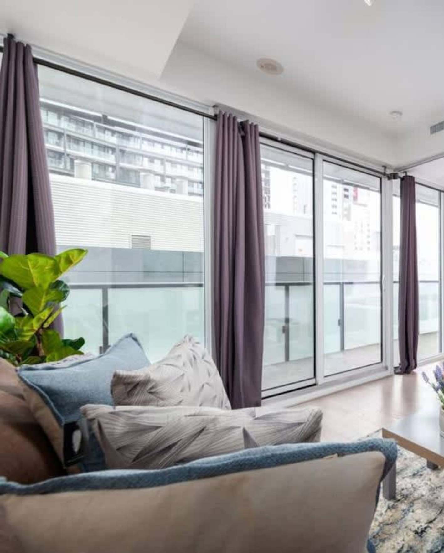 A bright high-rise living room with floor-to-ceiling sliding glass doors and a glass balcony railing overlooking neighboring apartment buildings. Mauve floor-length curtains frame the windows. In the foreground, a tan sofa with textured gray and blue accent pillows sits beside a large fiddle-leaf fig plant. A blue knit throw is draped over the armrest, and light hardwood flooring extends to the balcony doors. The white ceiling features recessed lighting and a soffit detail.