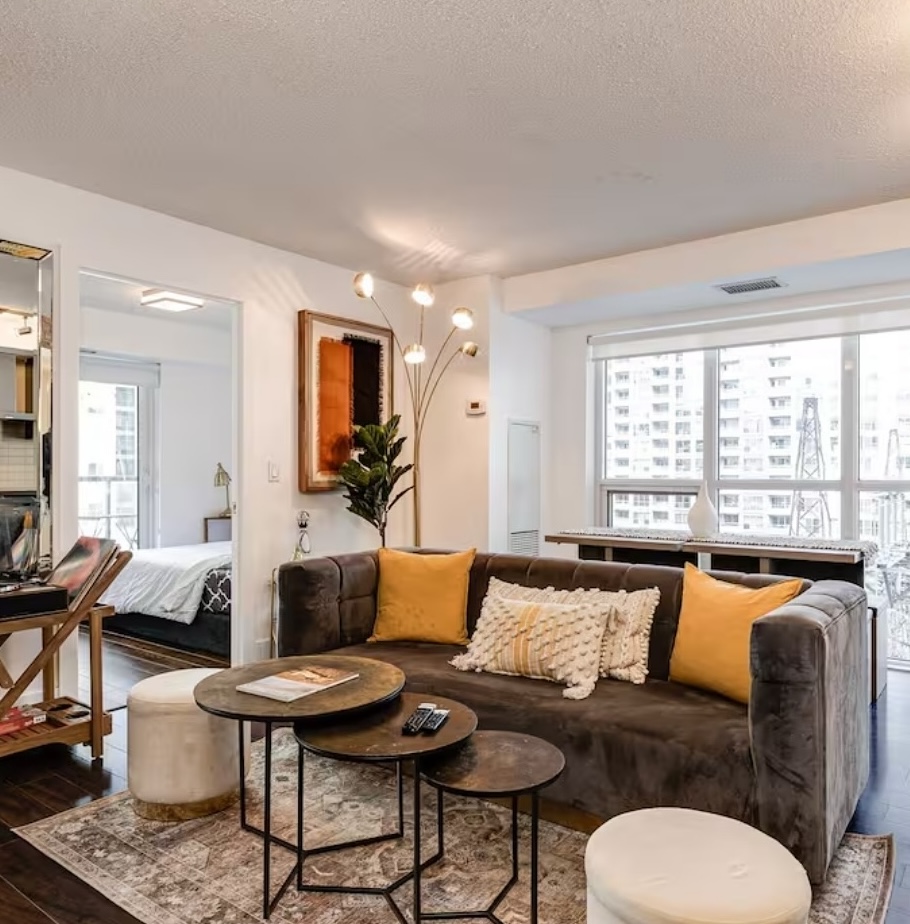 Modern Airbnb living room with velvet sofa, nesting coffee tables, accent lighting, large windows, and an open view into the bedroom.