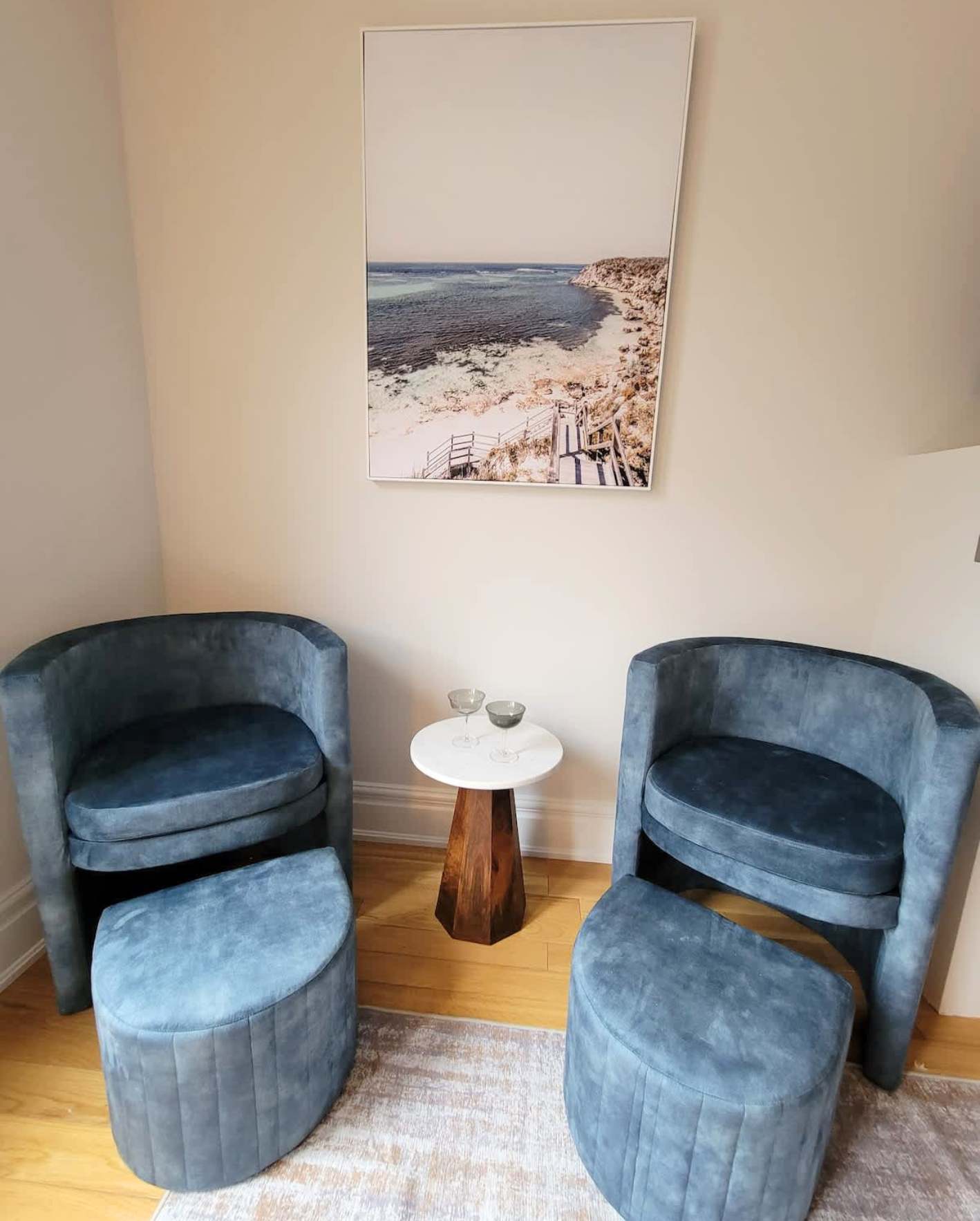 A cozy sitting nook with warm beige walls and honey hardwood flooring. Two dusty blue velvet barrel chairs with matching round channel-tufted ottomans face each other, separated by a small white marble-top side table with a geometric natural wood pedestal base. Two stemmed glasses sit on the table. Above, a large framed coastal photograph shows a sandy beach path with wooden stairs leading down to a calm turquoise shoreline and rocky cliffs. A light gray area rug partially covers the floor beneath the seating arrangement.