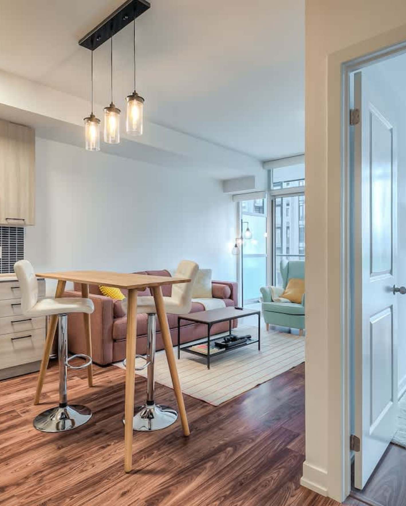 An open-concept high-rise apartment with warm walnut-toned hardwood flooring viewed from a hallway entrance. A tall wooden pub table with two white leather adjustable bar stools sits beneath a black track-mounted triple pendant light with glass cylinder shades. Beyond, a mauve sofa with a metal-frame coffee table sits on a cream area rug, facing floor-to-ceiling windows overlooking neighboring buildings. A sage green wingback armchair with a yellow accent pillow occupies the far corner. To the left, light wood kitchen cabinetry and a white countertop are partially visible.