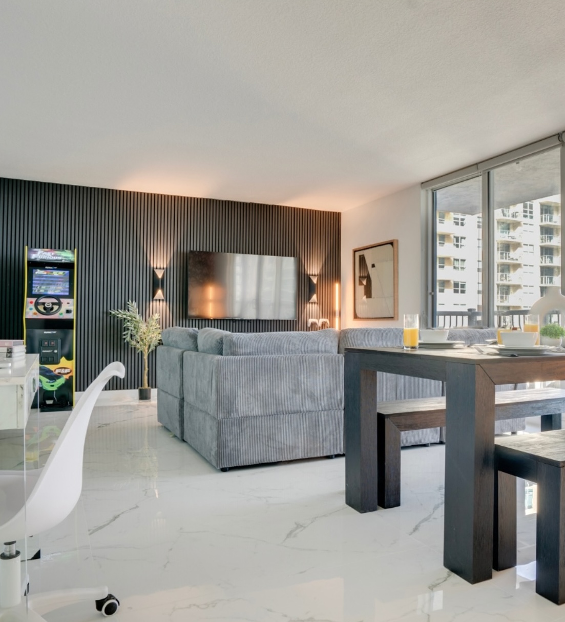 Modern Airbnb living and dining area with sectional sofa, wall-mounted TV, dark wood dining table, arcade game, and floor-to-ceiling balcony windows.